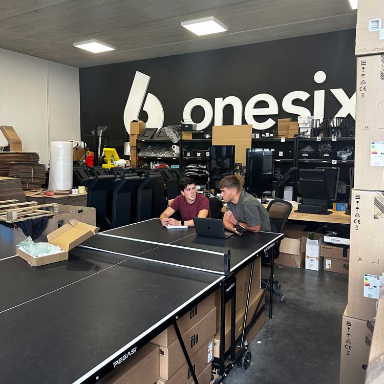 Two men sit at a ping pong table with a laptop in a warehouse filled with boxes and equipment, with a large 'onesix' logo on the wall behind them.