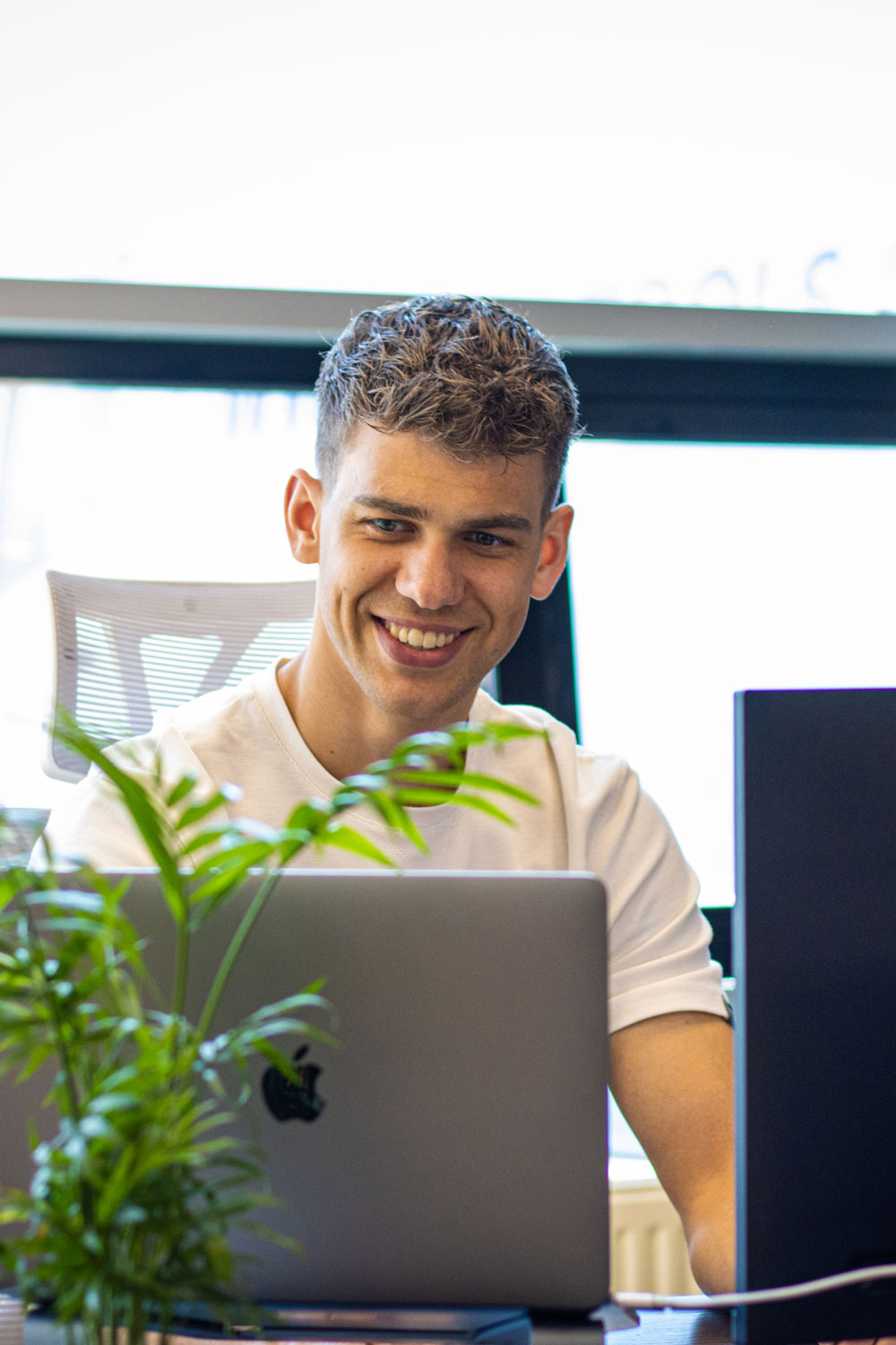 Smiling young man (Zoe Onesix) with short curly hair sitting at a desk working on a laptop in a bright office with a plant in the foreground.